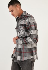 Cămașă din flanel în carouri gri și negre, cu accente roșii, buzunar pe piept butonat și o croială ușor relaxată. Textura pare moale și confortabilă.