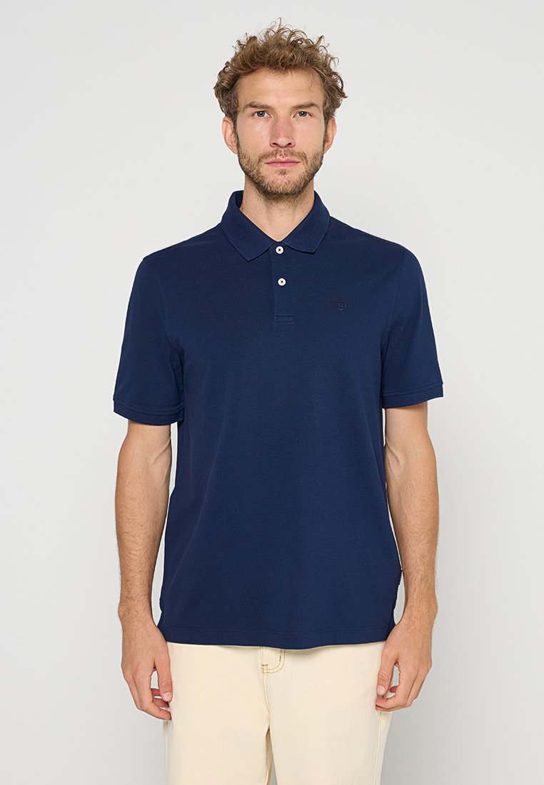 Homme aux cheveux bouclés portant un polo à manches courtes bleu marine et un pantalon beige clair, se tenant devant un fond clair uniforme.