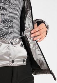 Silver pufferjacka med en glänsande, vadderad yttersida, svarta detaljer, dragkedja fram och knappstängning; insidan har en mönstrad design.