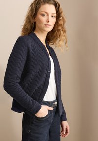 Veste matelassée marine avec un col rond, équipée de boutons-pression et d'un tissu texturé. Associée à un haut blanc et à un jean bleu marine.