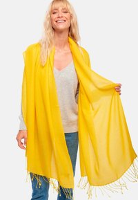 Femme souriante avec de longs cheveux blonds portant un grand châle frangé jaune vif par-dessus un pull gris clair et un jean bleu.