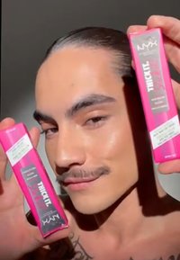 Due tubetti di mascara rosa con testo nero, tenuti da una persona con baffi. Il prodotto è etichettato "THICK IT" e presenta una finitura lucida.