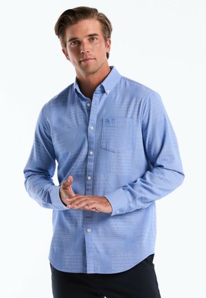 Man draagt een lichtblauw overhemd met patroon en borstzak, staat met gevouwen handen tegen een witte achtergrond.