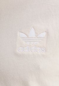 Krämfärgad tyg med en broderad vit Adidas-logotyp och tre bladliknande streck ovanför varumärkesnamnet i gemener.