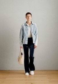 Persona stāv iekštelpās, ģērbusies gaišā džinsu jakā, baltā kreklā, melnās biksēs, baltos sporta apavos un tur rokās bēša krāsas rokassomu.