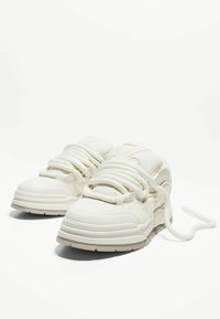 Baskets en cuir blanc avec un design courbé, semelle épaisse et proéminente, accents texturés et lacets épais, présentant une finition lisse et des détails discrets.