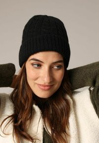 Zwarte geribbelde gebreide beanie met een textuurpatroon. Gemaakt door een persoon met lang bruin haar, gedragen in combinatie met een groene trui en een crèmekleurige buitenjas.