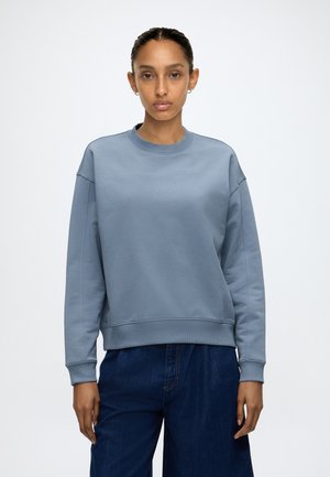 Femme portant un sweat-shirt bleu à col rond et un jean bleu foncé, debout sur un fond blanc uni.