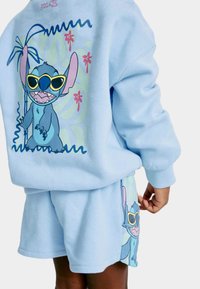 Světle modrý bavlněný sweatshirt a šortky se vzorem modré postavy v brýlích na pozadí tropické krajiny.