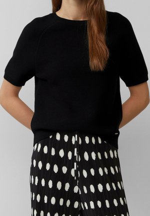 Frau trägt ein schwarzes Kurzarmoberteil, in einen schwarzen, plissierten Rock mit weißen Punkten gesteckt, steht mit den Händen hinter dem Rücken.