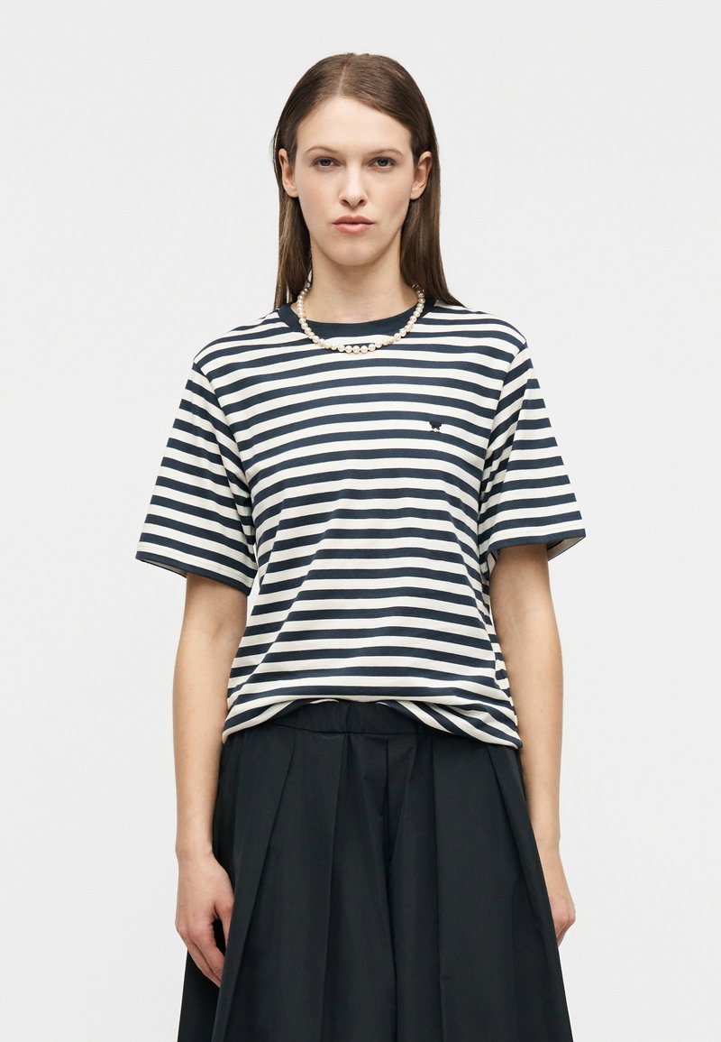 Γυναίκα με καστανά ίσια μαλλιά, φορεί ένα μαύρο και λευκό ριγέ μπλουζάκι, μαύρη πλισέ φούστα και μαργαριταρένιο κολιέ, μπροστά σε απλό φόντο.