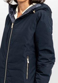 Giacca a vento blu navy con cappuccio e zip dorate, dotata di una fodera trapuntata e due tasche laterali. Texture morbida e leggera coibentazione.