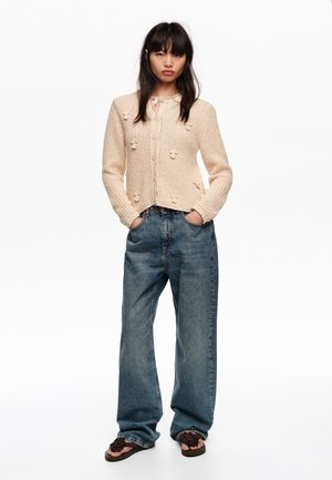 Kobieta ubrana w beżowy sweter z dzianiny z teksturowanymi detalami, luźne niebieskie dżinsy i brązowe sandały z plecionki, stojąca z rękami w kieszeniach.