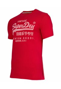 Rode T-shirt met korte mouwen, gemaakt van katoen, met een ronde hals. Bevat witte tekst met het "SuperDry"-merkimago en andere graphics.
