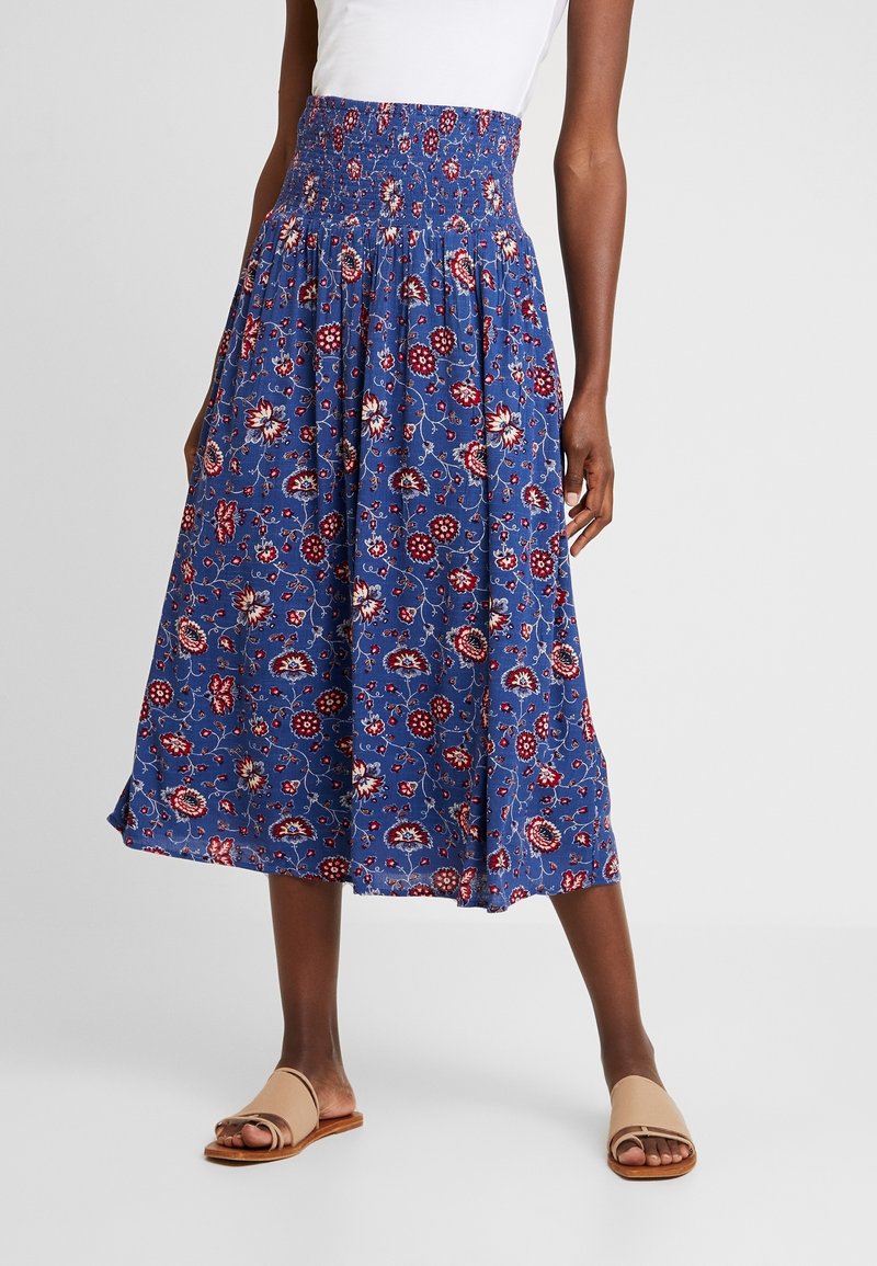 Blaue Midirock mit Blumenmuster in Rot und Weiß, mit einem geschoppten hohen Bund und fließendem Design. Getragen mit beigen Rutschsandalen.