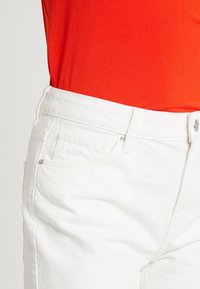 Jeans de mezclilla blanca con cinco bolsillos, un botón delantero y trabillas para cinturón. Combinados con una blusa roja brillante, que muestra una textura suave.