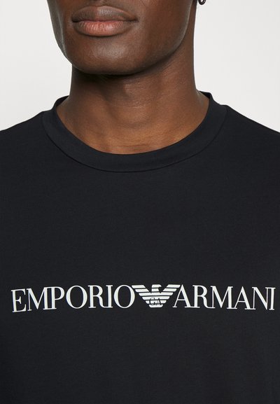 Juodas medvilnės marškinėlis su apvaliu kaklu ir baltu Emporio Armani logotipu su sparnuotu simboliu per krūtinę. Lygus paviršius.