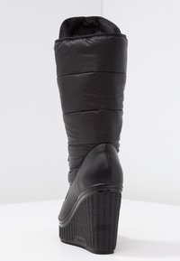 Bota de cuña negra con parte superior acolchada y acolchada, y detalles de cuero liso. La cuña es texturizada, con un diseño acanalado y marca.