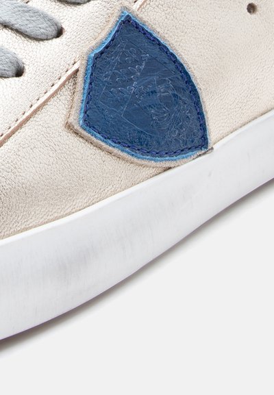 Ανοιχτό μπεζ αθλητικό παπούτσι με επάνω μέρος από δερμάτινο υλικό με υφή, γκρι κορδόνια και μπλε δερμάτινο έμπλαστρο με ανάγλυφες λεπτομέρειες. Λευκή σόλα από καουτσούκ.