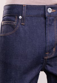 Tmavomodré džíny z denimu mají oranžové švy, kovový knoflík s logem a přední kapsu na levé straně.