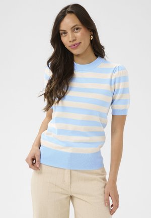 Młoda kobieta z długimi brązowymi włosami, ubrana w niebiesko-biały sweter w paski z krótkim rękawem i beżowe spodnie, stojąca na jednolitym tle.