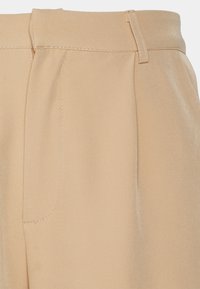 Calças de tecido bege com uma textura suave, frente plissada e uma cintura com fecho de gancho e barra. Bolso lateral visível.