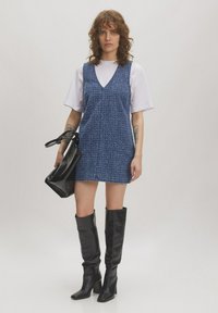 Denim mini-jurk van een gestructureerde blauwe stof, met een diepe V-hals, gecombineerd met een wit T-shirt en hoge zwarte leren laarzen. Zwarte handtas.