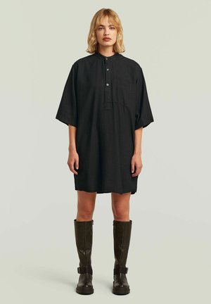 Kobieta z blond włosami ubrana w luźną czarną sukienkę koszulową z guzikami i kieszenią na piersi, połączoną z ciemnobrązowymi kozakami za kolano.