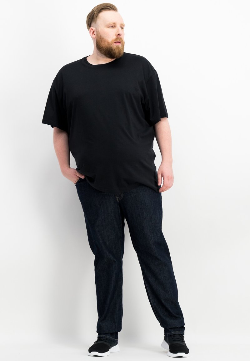 Maglietta nera a maniche corte in morbido tessuto, abbinata a jeans blu scuro e sneakers nere, in piedi su uno sfondo bianco.