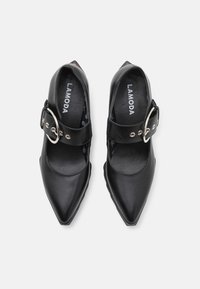 Zapatos de cuero negro con punta afilada, con una correa ancha y una hebilla metálica circular. El interior presenta un patrón sutil con la marca.