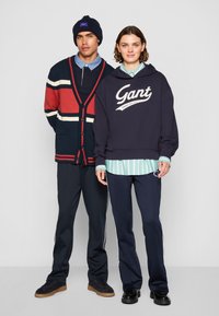 Deux modèles se tiennent ensemble. Le modèle masculin porte un cardigan rayé bleu marine, rouge et blanc par-dessus une chemise à col, ainsi que des pantalons bleu marine. Le modèle féminin porte un hoodie bleu marine avec le logo "Gant", superposé sur une chemise rayée teal et un pantalon bleu marine.