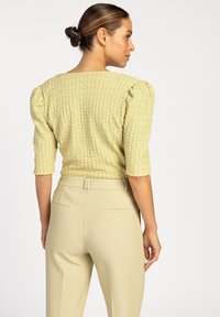 Geel getextureerde top met pofmouwen, gecombineerd met lichtbeige op maat gemaakte broek. De stof heeft een subtiel verhoogd patroon.