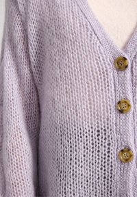 Cardigan lavorato a maglia di un tenue viola con scollo a V, caratterizzato da tre bottoni rotondi marroni, una textura morbida e leggera con una leggera lucentezza.