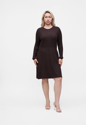 Femme aux longs cheveux blonds portant une robe en maille à manches longues marron foncé et des sandales à talons beiges, debout sur fond blanc.