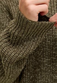 Maglione marrone lavorato a maglia con polsini a costine, caratterizzato da un motivo testurizzato e lievi sfumature di colore chiaro in tutta la stoffa. Inquadratura ravvicinata del maniche.