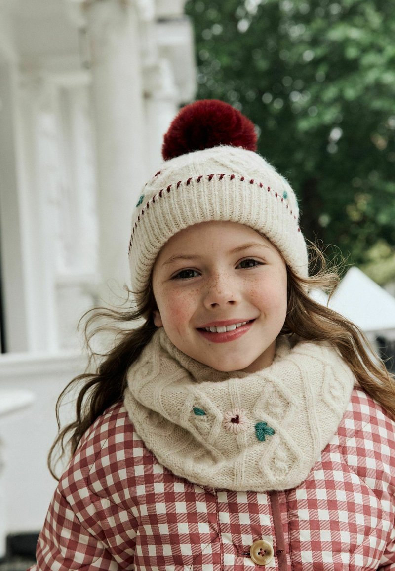 Enfant souriant portant un bonnet tricoté crème avec un pompon rouge, une écharpe assortie et une veste rouge à carreaux dehors avec des colonnes blanches et des arbres verts.
