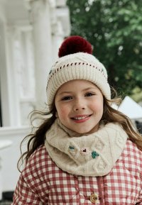 Strik hvid hue med rød pom-pom, røde og grønne pynt; creme kabelstrikket tørklæde med blomsterakcenter; maroon ternet quiltet frakke.