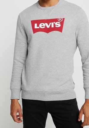 Homme portant un sweat-shirt Levi's gris clair avec un logo rouge et blanc sur la poitrine, associé à un pantalon noir.