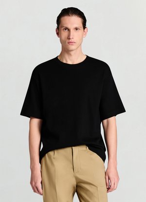 Schwarzes T-Shirt mit kurzen Ärmeln und rundem Ausschnitt, aus weichem Stoff. Es hat eine lockere Passform und eine glatte Textur, kombiniert mit beigen Hosen.