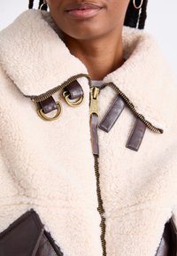 Giacca in finta pelliccia shearling color crema con colletto in pelle marrone, dotata di una cerniera dorata, dettagli in doppio anello D e pannelli di tessuto marrone scuro.