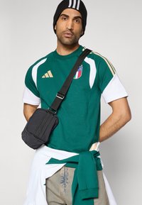 Grøn og hvid atletisk skjorte med gulddetaljer og italiensk våbenskjold, parret med cowboybukser og en sort skuldertaske. Sort beanie med striber.