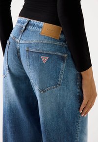Vaqueros de mezclilla de color azul claro con un diseño de pierna ancha, presentando un parche de piel marrón en la cintura trasera y un logo triangular en el bolsillo derecho.