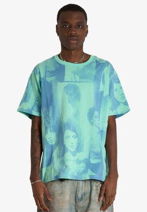 Giovane uomo che indossa una maglietta turchese larga con stampe sbiadite in bianco e nero di ritratti e jeans blu sbiaditi, in piedi davanti a uno sfondo bianco.