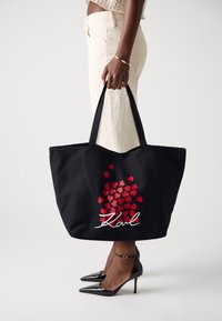 Sac fourre-tout noir avec des motifs de cœurs rouges et le texte blanc "Karl". Matériau en toile, forme rectangulaire, doubles poignées. Chaussures à talons hauts noires dans le cadre.