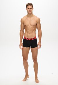 Boxer neri con un bordo rosso brandizzato "SUPERDRY"; il modello è a piedi nudi, in piedi con le braccia lungo i fianchi, su uno sfondo neutro.