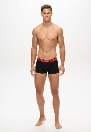 Boxer shorts noirs avec une ceinture de marque "SUPERDRY" rouge ; le modèle est pieds nus, debout avec les bras le long du corps, sur un fond neutre.