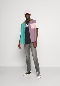 Multicolored short-sleeve button-up shirt with teal, purple, and cream sections, featuring a chest pocket and worn with gray jeans and black sneakers.