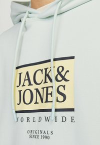 Jasnoniebieska bluza z kapturem z dużym żółto-czarnym nadrukiem graficznym. Posiada sznurki ściągające oraz miękką fakturę. Tekst zawiera "JACK & JONES WORLDWIDE."