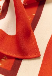 Rød og cremefarvet mønstret stof med en glat overflade. Har en foldet kant og synlige syninger, der fremhæver geometriske design.