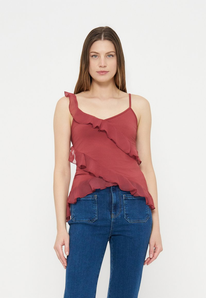 Femme portant un haut sans manches rose foncé à volants avec fines bretelles et un jean bleu, debout devant un fond blanc uni.